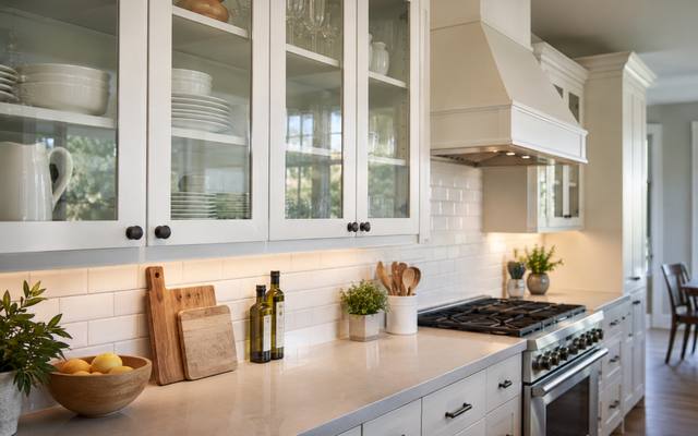 Glass-front uppers and a range hood in a bright white compact kitchen.